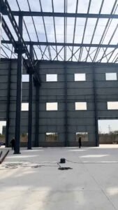Steel frame of a logistics warehouse building showing large wall openings for ventilation, with a pickup truck outside.
