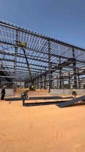 Trabajadores instalando paneles de cubierta en una nave industrial prefabricada de acero con asistencia de maquinaria elevadora.