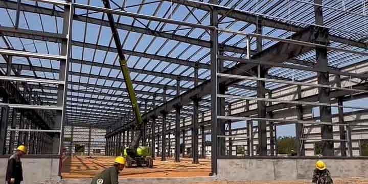 Construcción de una nave industrial prefabricada con grúa instalando vigas de acero mientras obreros con cascos amarillos trabajan en la obra bajo un cielo soleado