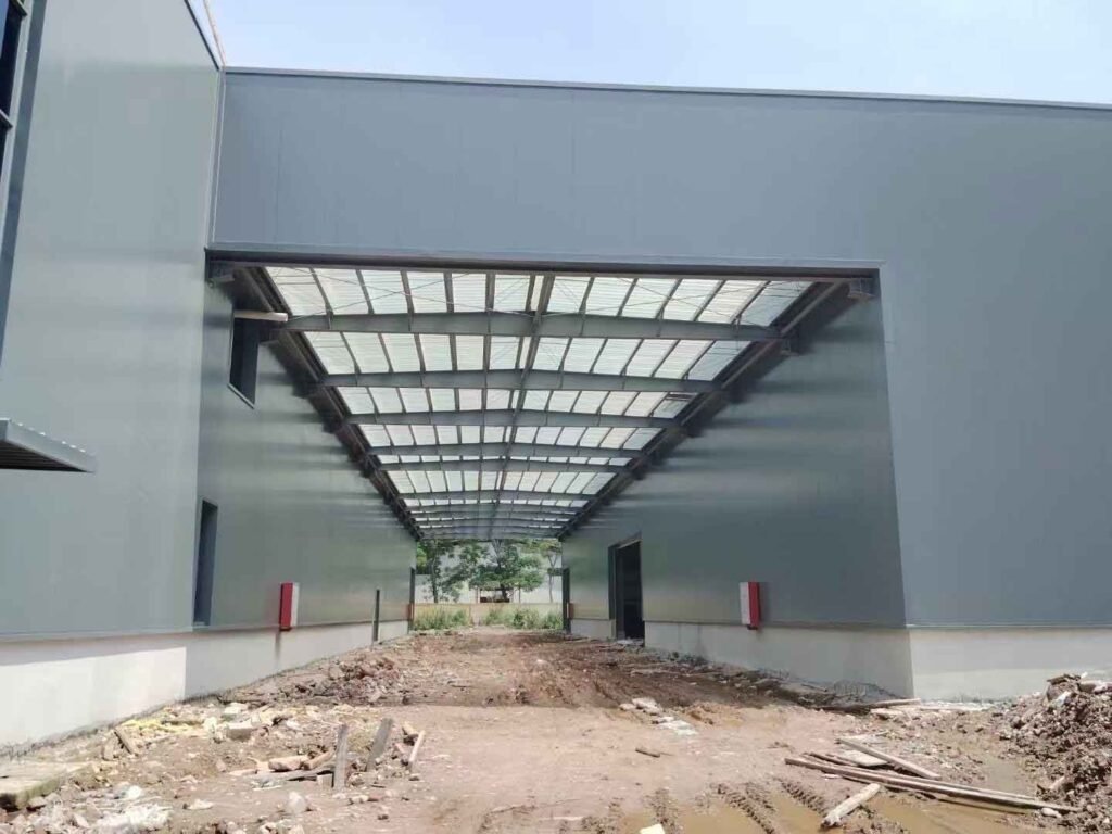 Amplio corredor de logística y área de carga entre almacenes industriales con techo de paneles traslúcidos y paredes laterales de chapa metálica gris.