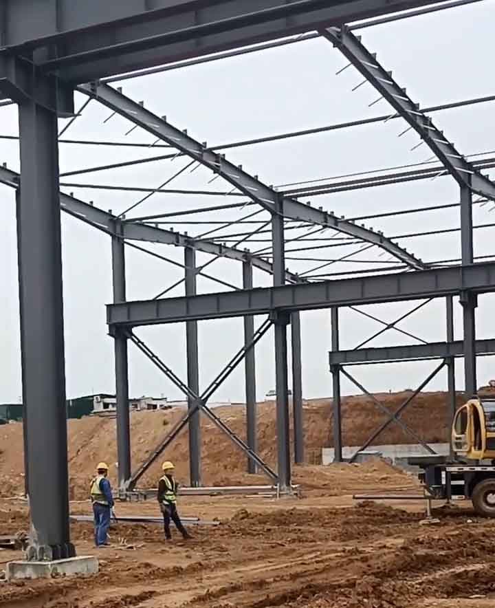 Arriostramientos en forma de X entre gruesas columnas grises de almacenes metálicos, con dos obreros supervisando el área frente a un montículo de tierra.