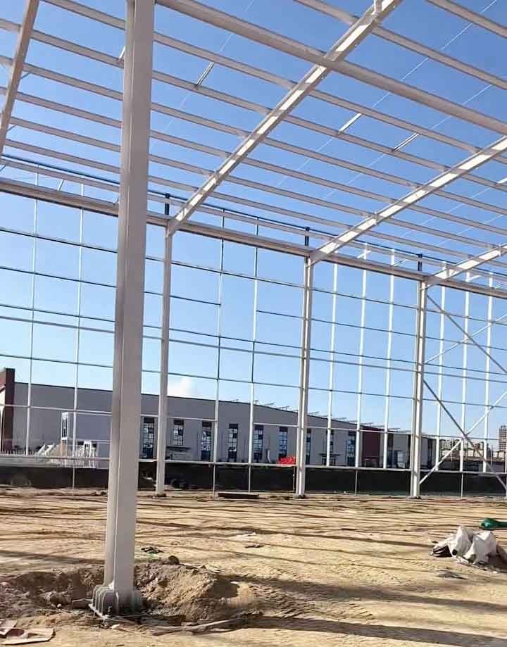 Detalle cercano de la base de una columna de acero blanco firmemente atornillada al cimiento en una obra de steel structure building, con vista hacia otros edificios al fondo a través de la perfilería.
