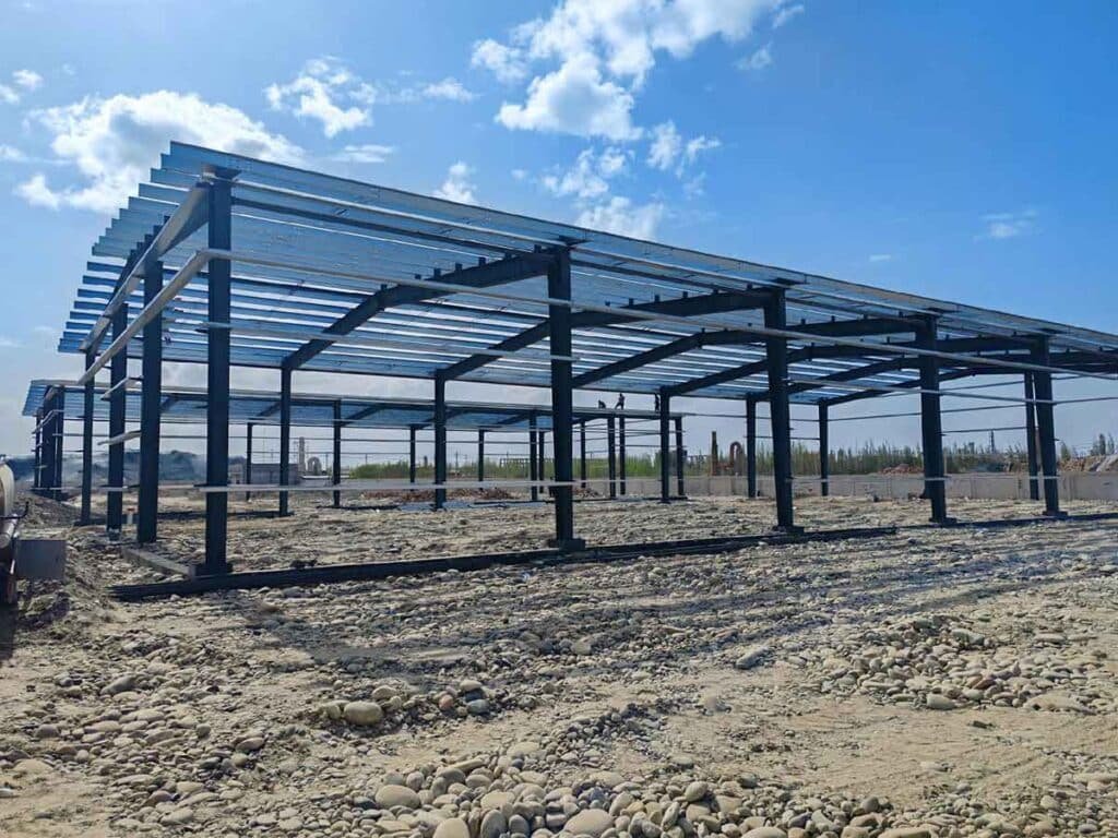 Black heavy-duty steel columns supporting partially installed light blue roof panels over a cobblestone terrain, reflecting an optimized steel warehouse price Colombia.