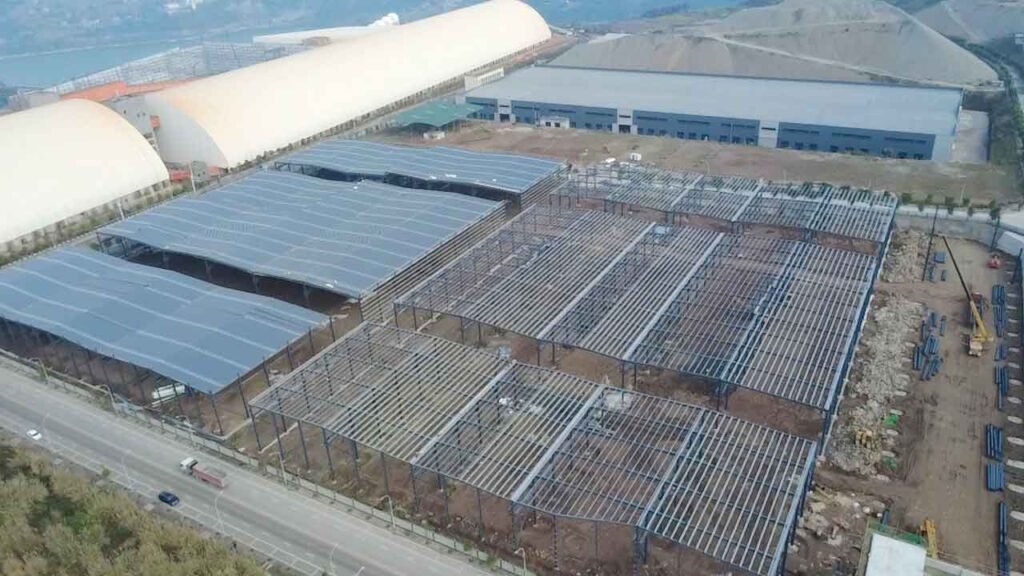 Vista aérea de una gran bodega industrial en construcción junto a una carretera, mostrando la mitad con cubierta instalada y la otra con armazón expuesto