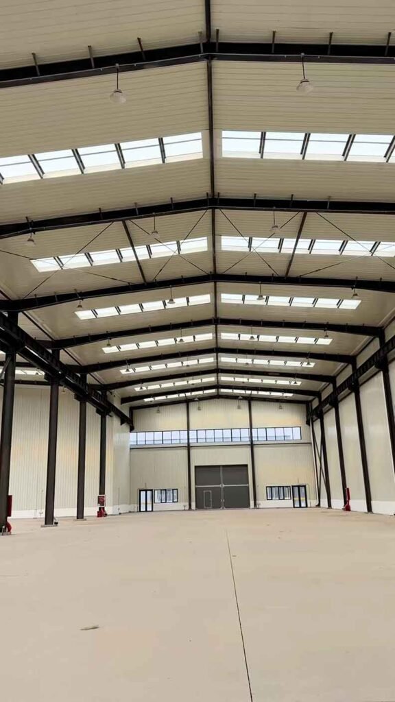 Interior de bodegas industriales de gran altura, destacando la estructura de acero negro, paredes de paneles aislantes blancos, filas de tragaluces en el techo y un gran portón corredizo gris al fondo con puertas peatonales.