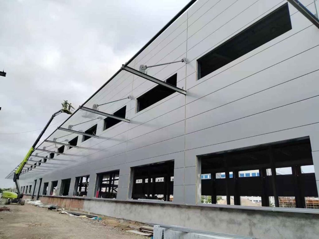 Operarios utilizando una plataforma elevadora verde para instalar con precisión paneles sándwich grises y soportes diagonales para marquesinas en la fachada de bodegas industriales prefabricadas.