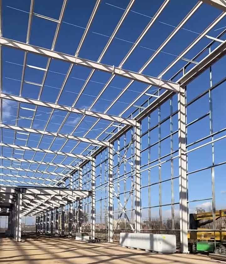 Entramado de techo de vigas blancas sin cubrir mostrando el cielo azul, característico de bodegas prefabricadas, con parte de un elevador articulado amarillo a la derecha