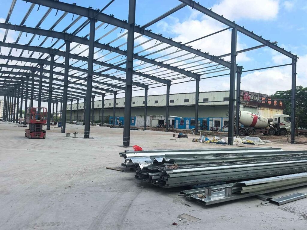 Sitio activo de construcción de galpones con un camión hormigonera trabajando al exterior y paneles de acero apilados en el interior.