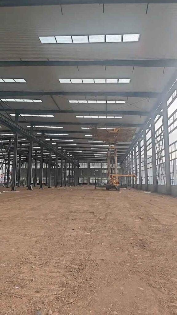 Plataforma de tijera amarilla operando bajo el techo con tragaluces durante la construcción de bodegas de acero gris sobre suelo de tierra.