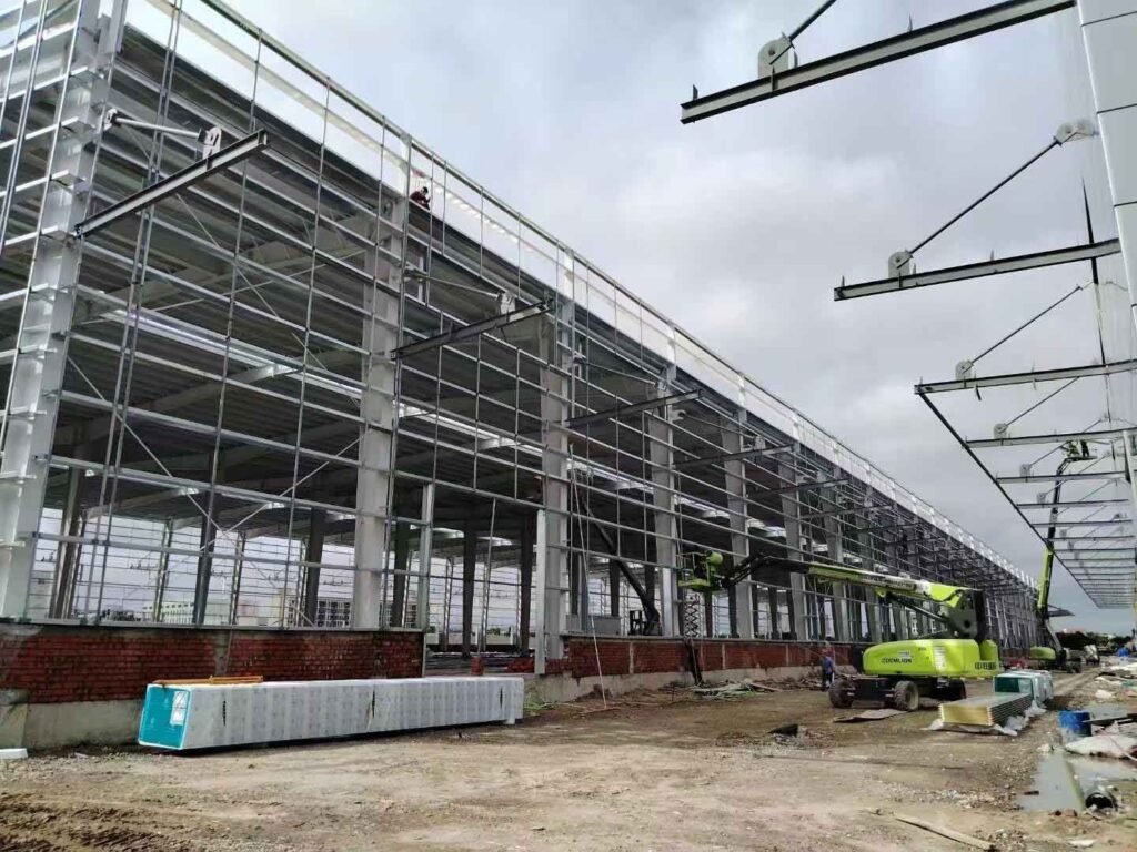 Dos plataformas elevadoras verdes operando dentro de una densa red de esqueleto principal de vigas H durante los trabajos de construcción de bodegas.