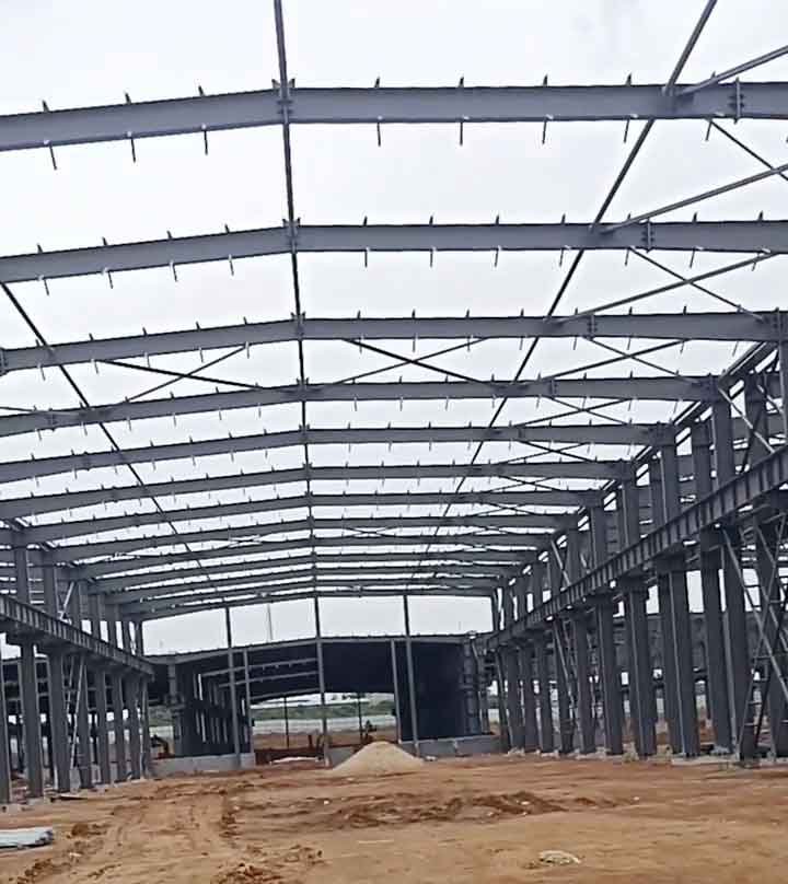 Vista interior profunda de galpones metálicos en construcción, mostrando vigas de acero gris oscuro con conectores superiores sobre un amplio piso de tierra rojiza.