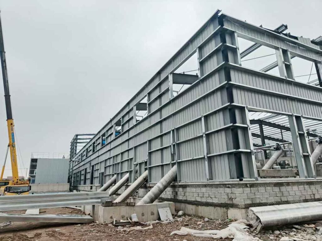 Montaje de estructura metálica industrial con columnas de acero, revestimiento parcial de chapa gris y una grúa amarilla operando en el fondo.