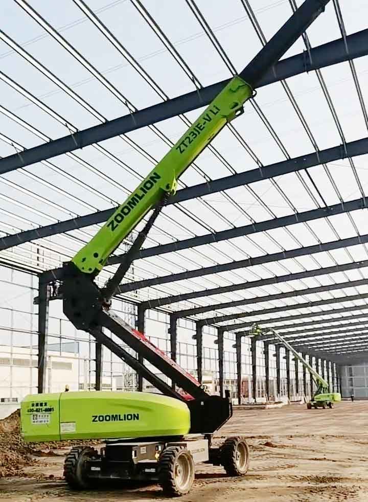 Plataforma elevadora articulada de color verde extendiendo su brazo hacia las vigas del techo durante una obra de construcción industrial.