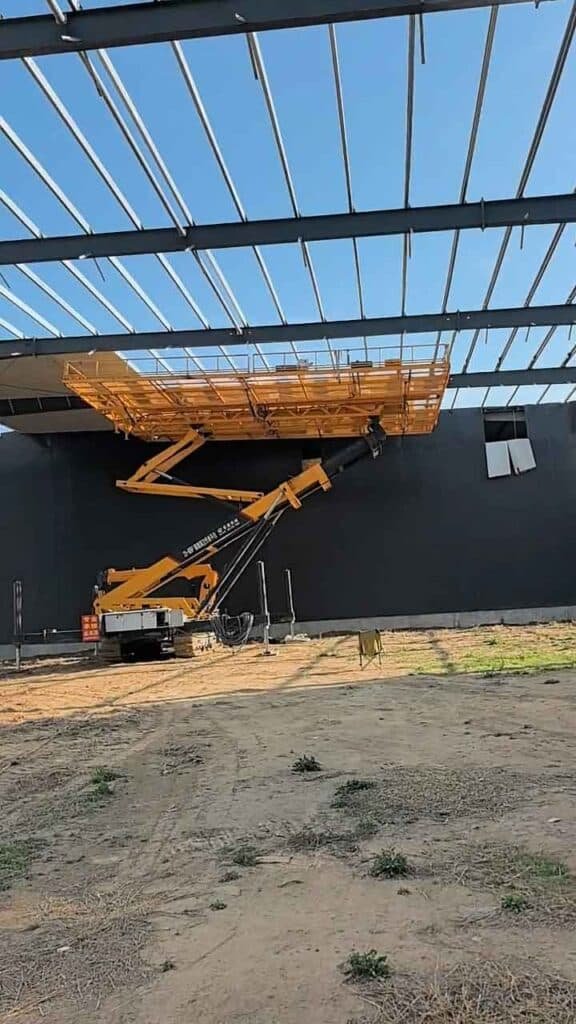 Plataforma elevadora amarilla de gran tamaño realizando el ensamblaje de estructuras bajo el techo de acero frente a una pared negra.