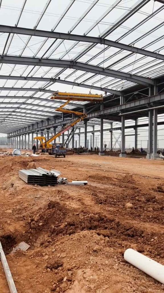 Ensamblaje estructural de columnas de acero negro ancladas a zapatas de concreto, junto a una zanja perimetral y un camión azul al fondo.