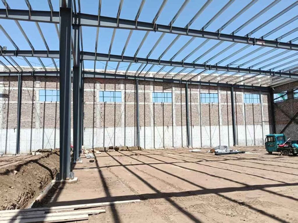 Ensamblaje estructural de columnas de acero negro ancladas a zapatas de concreto, junto a una zanja perimetral y un camión azul al fondo.