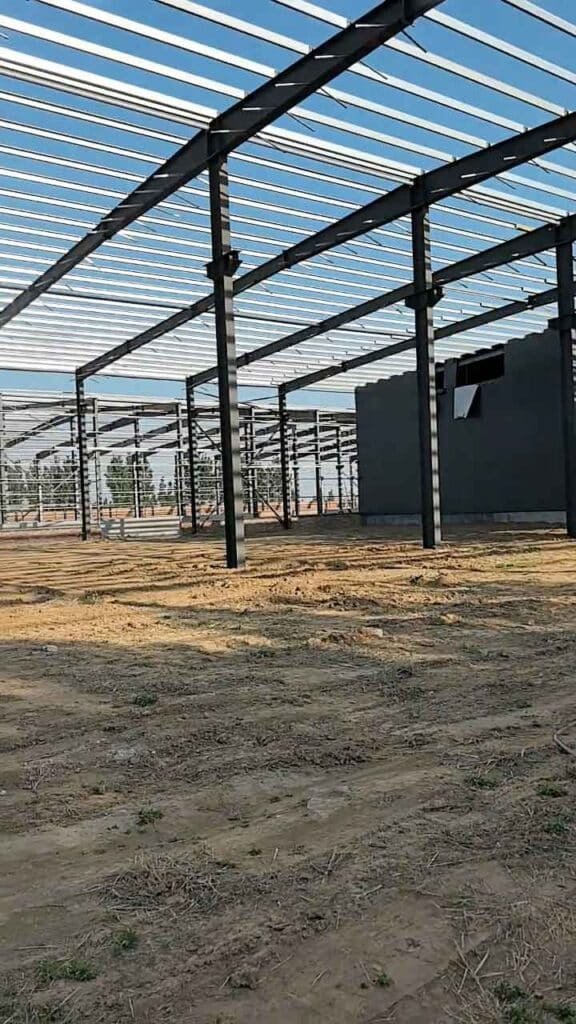 Filas de robustas estructuras de acero negro formando el esqueleto de una nave industrial con suelo de tierra y una pared lateral oscura a la derecha.