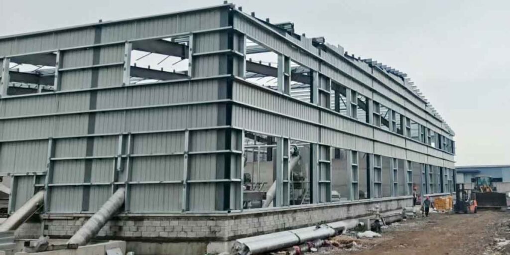Vista amplia de un gran almacén industrial mostrando una estructura de acero de dos niveles, paredes parcialmente revestidas y maquinaria pesada en el exterior.