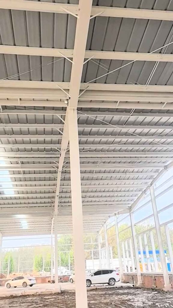 Vista ascendente de una columna blanca que sostiene la cubierta de una estructura industrial metálica, con tensores visibles y dos coches aparcados en el exterior al fondo.