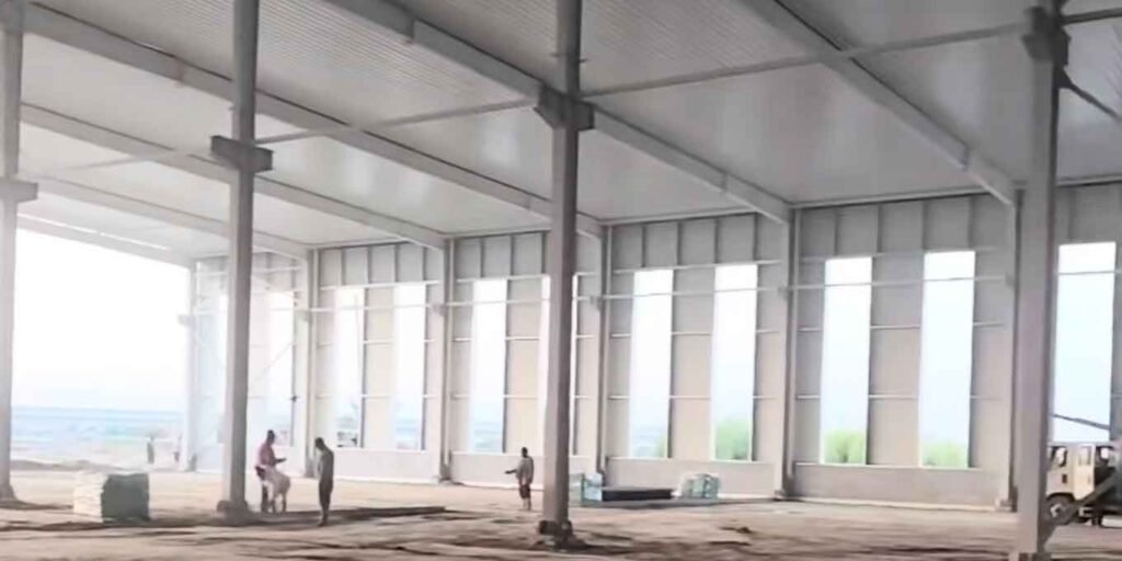 Vista interior de una estructura metálica prefabricada de color blanco con la cubierta superior totalmente instalada, grandes vanos en las paredes y un grupo de trabajadores sobre el piso de tierra.