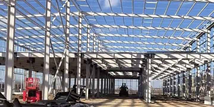 Amplia vista de estructuras metálicas pintadas de blanco formando el esqueleto de un edificio, con un nivel de entrepiso a la derecha y una plataforma elevadora roja sobre el suelo de tierra.