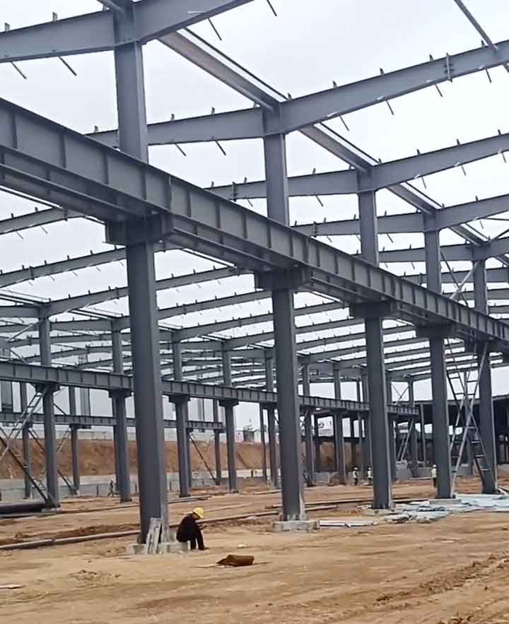 Entramado de estructuras metálicas prefabricadas de color gris, con un trabajador de casco amarillo sentado en la base de hormigón de una columna principal.
