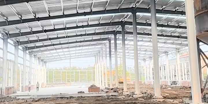 Vista interior amplia de estructuras metálicas prefabricadas pintadas de blanco, mostrando techo de chapa corrugada con tragaluces y dos personas a lo lejos sobre piso de tierra.