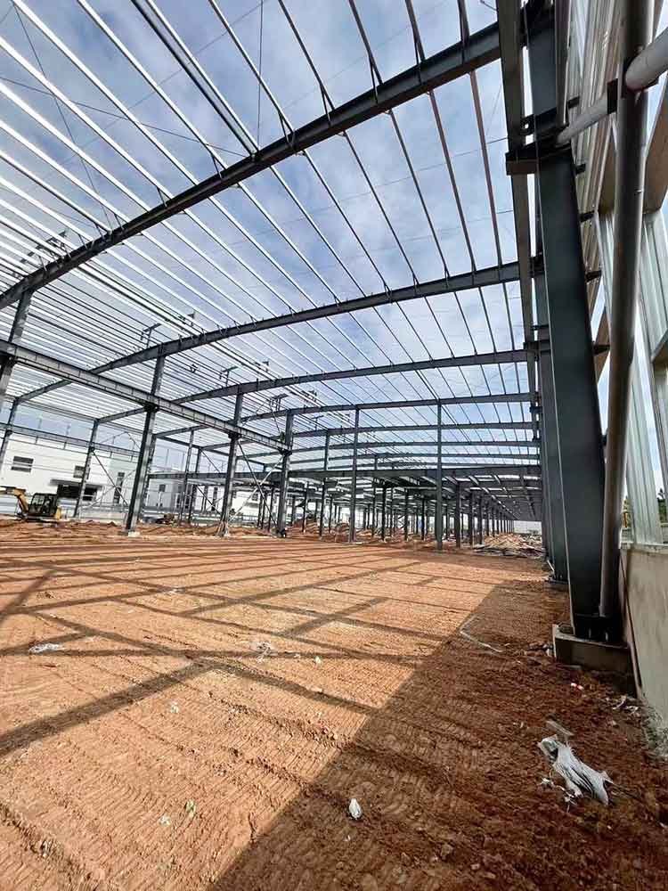 Interior perspective showing robust H-beam columns and grid-like roof shadows on the red dirt floor of a large clear-span steel structure building Ecuador