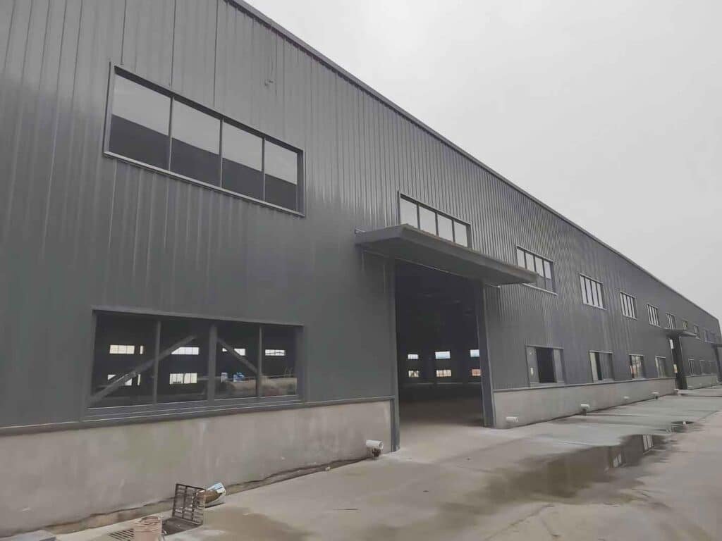 Fachada exterior de una fabrica de galpones metalicos con recubrimiento de lámina gris, ventanas laterales y marquesina reforzada para accesos logísticos