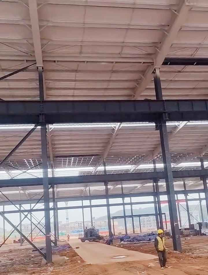 Un obrero con casco amarillo camina por el interior de una gran instalación de fabricación industrial mostrando fuertes vigas horizontales oscuras.