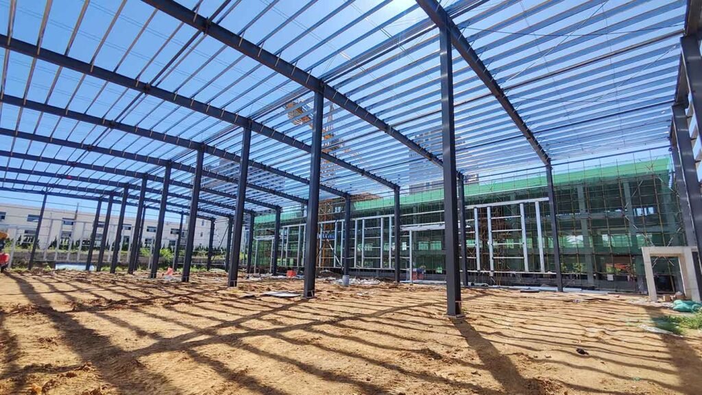 Amplio espacio interior de galpones de acero chinos en construcción, con vista a una plataforma elevadora roja y el edificio adyacente cubierto con andamiaje de seguridad verde.