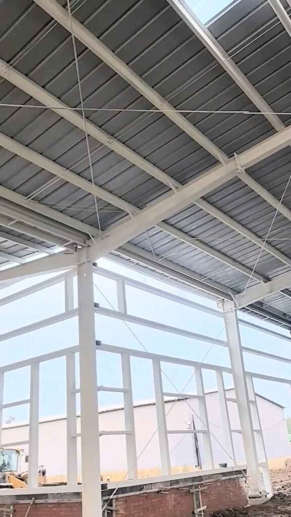 Entramado lateral para cerramientos en galpones metálicos de color blanco, apoyado sobre un pequeño muro de ladrillo, con una excavadora amarilla asomando por la izquierda.