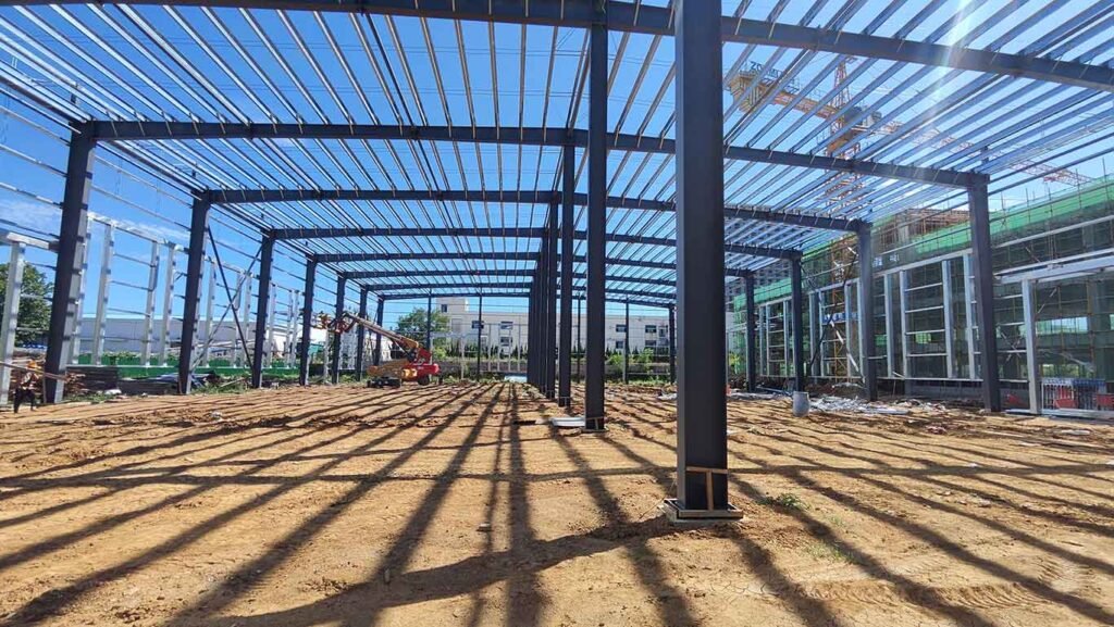 Columnas pesadas y entramado de techo de galpones metálicos chinos en fase de instalación, con un destello de sol brillante y vistas a un edificio con andamios verdes a la derecha.