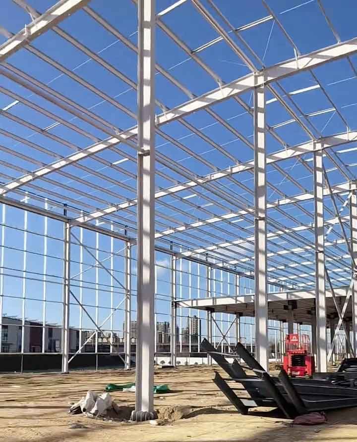 Pilares y vigas blancas de galpones metálicos con piezas de acero oscuro apiladas en el suelo de arcilla en primer plano y una plataforma elevadora roja trabajando al fondo.