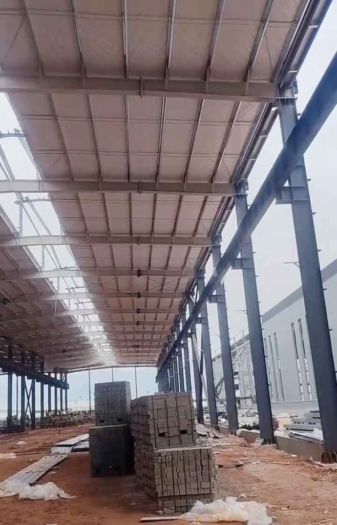 Pilas de bloques de hormigón gris almacenadas bajo la cubierta clara y pilares oscuros durante el proceso constructivo de galpones metálicos.