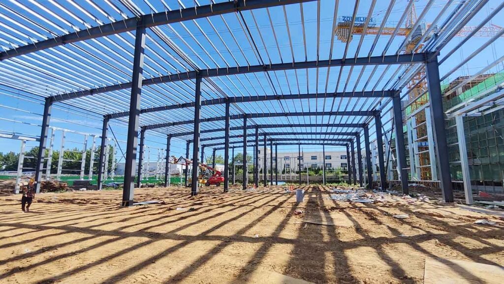 Estructura de acero bajo construcción para galpones prefabricados chinos, mostrando sombras lineales de las vigas en el suelo de tierra, un trabajador a la izquierda y una grúa torre amarilla al fondo.
