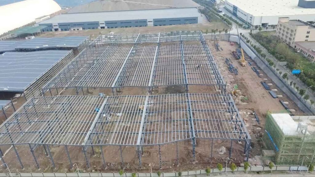 Vista aérea de la construcción de una nave industrial donde una grúa móvil amarilla opera junto a perfiles azules apilados sobre el terreno.