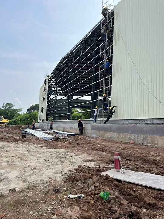 Operarios trabajando en andamios verticales altos instalando paneles de chapa ondulada clara en la fachada exterior de un galpón prefabricado