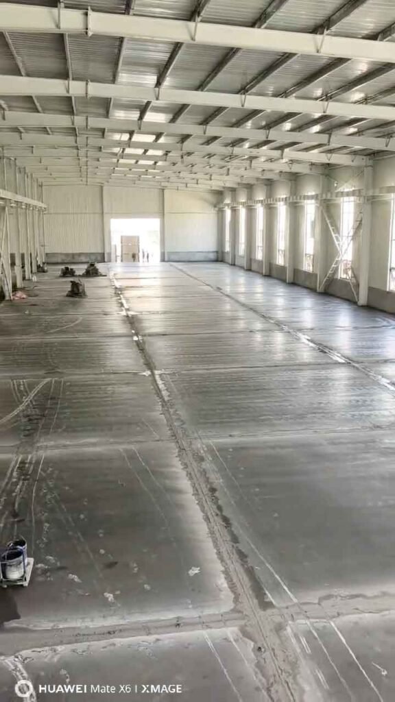 Vista longitudinal del piso de concreto gris con cortes de expansión en el amplio interior de galpones industriales de estructura metálica blanca.