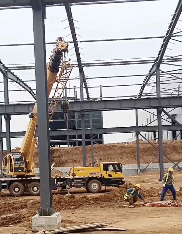 Una grúa móvil amarilla elevando componentes de acero gris mientras obreros coordinan la maniobra en el suelo de tierra para bodegas industriales.