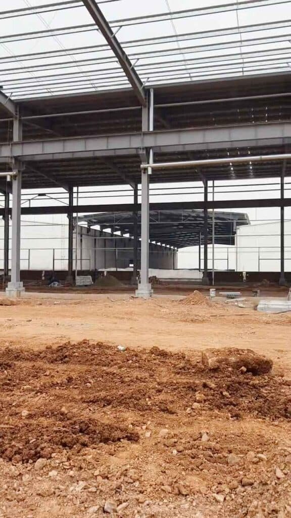 Nave en construcción mostrando robustas estructuras de acero, un piso de tierra con sombras lineales y un gran hueco en la pared por donde se ve una excavadora amarilla.