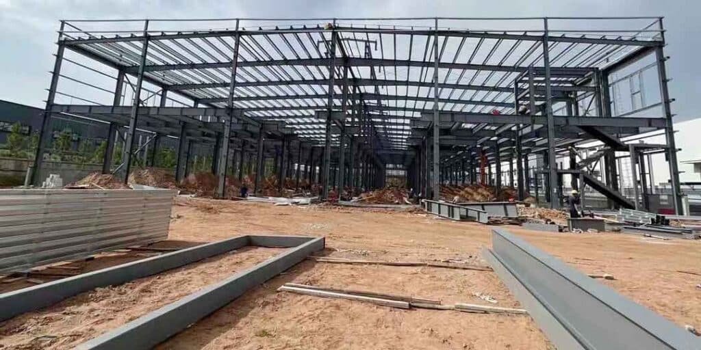An active job site showing stacks of metal decking materials and loose beams in the foreground of a massive multi-story framework during heavy steel structure construction.