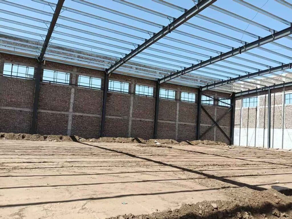 Interior de nave demostrando una construcción eficiente con muros de ladrillo rojo, ventanas superiores y tensores de acero en cruz en la estructura de soporte.