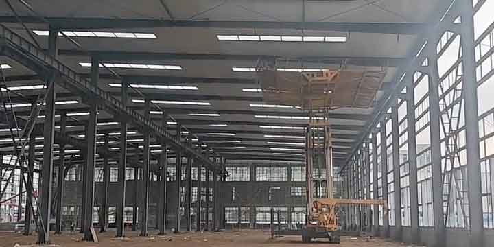 Plataforma elevadora amarilla trabajando directamente bajo el techo de paneles blancos con franjas transparentes de un amplio steel warehouse en construcción.