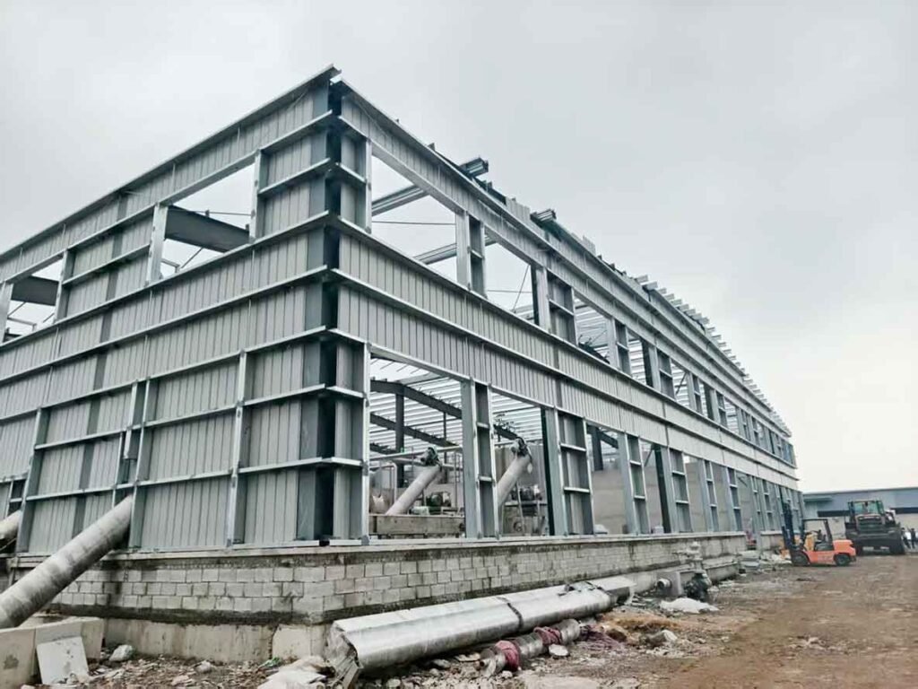 Esqueleto de acero expuesto de un galpón industrial en construcción, con marcos para ventanas, tubos de soporte diagonal y un montacargas operando.