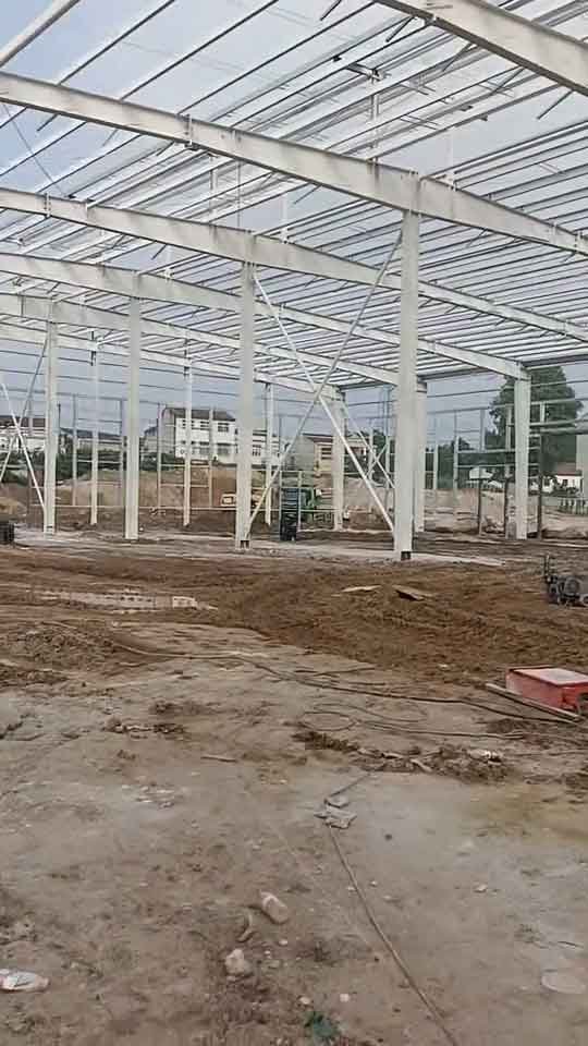 Dos plataformas elevadoras amarillas operando para ensamblar una nave industrial prefabricada blanca sobre un terreno muy fangoso con grandes charcos de agua.