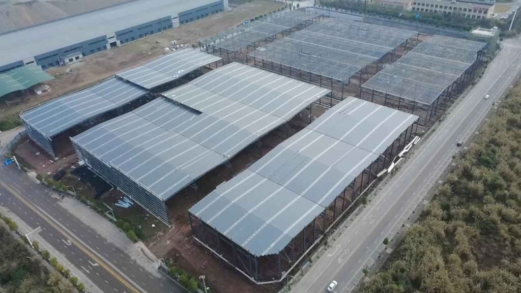 Complejo logístico de múltiples módulos mostrando su estructura metálica de techo y cubierta gris, situado en paralelo a una avenida arbolada.