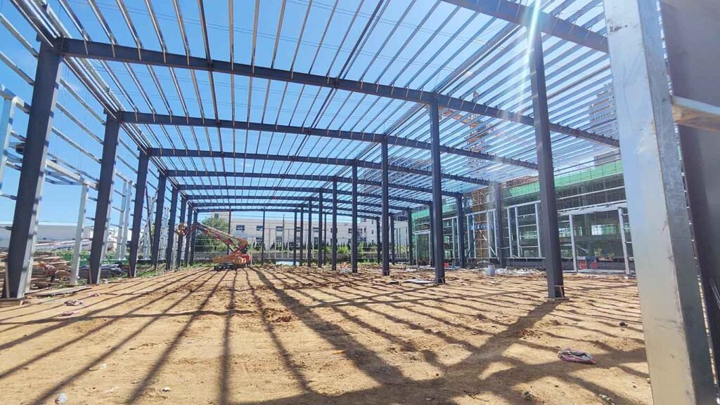 Primer plano de una columna de acero estructural masiva en la obra de naves industriales chinas, con fuerte iluminación solar filtrándose por el techo sin cubrir y maquinaria trabajando al fondo.