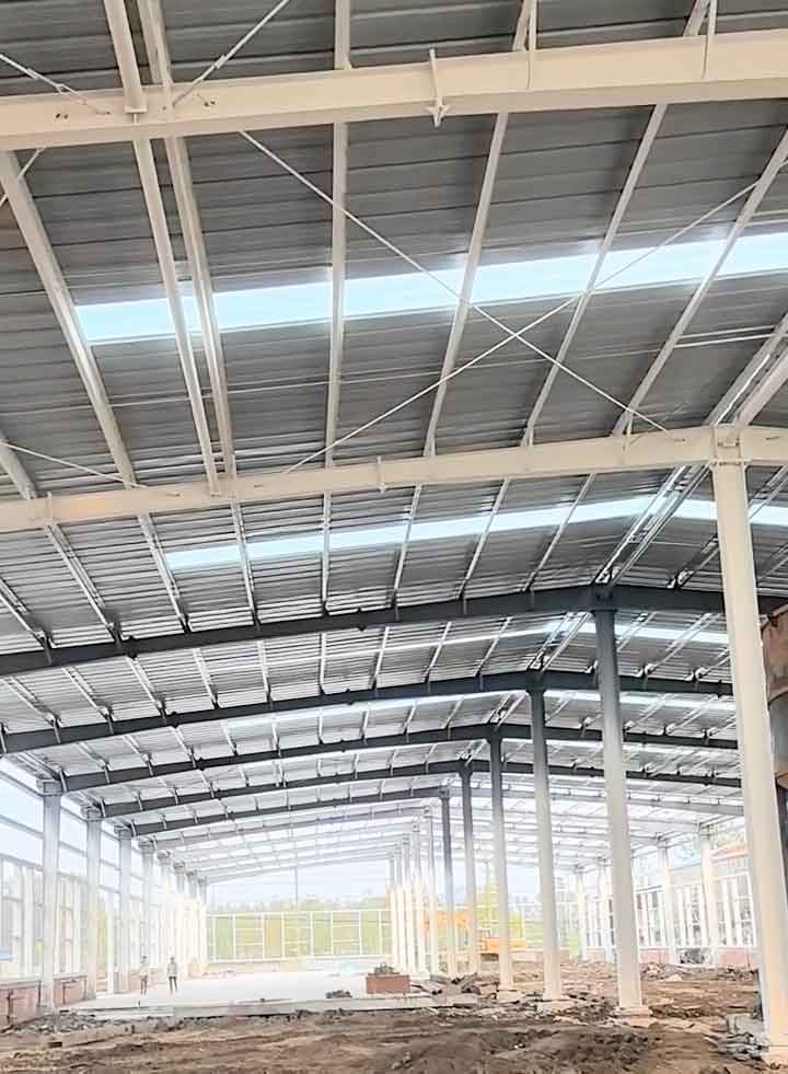 Profundidad del armazón de acero blanco de naves industriales, resaltando la cubierta ondulada brillante y el zócalo perimetral de ladrillo rojo sobre el suelo fangoso.