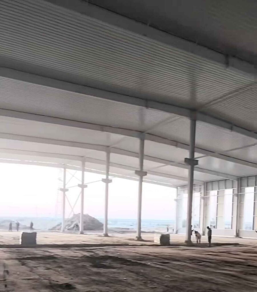 Gran espacio diáfano característico de naves industriales, mostrando techo cerrado de paneles blancos, gruesas columnas alineadas y bloques de construcción apilados sobre el suelo de tierra.